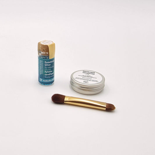 Beauty-Set mit Shea-Butter, Auftragspinsel und intensiv metallblauem Bio-Glitzer in Glasflasche auf hellem Untergrund.