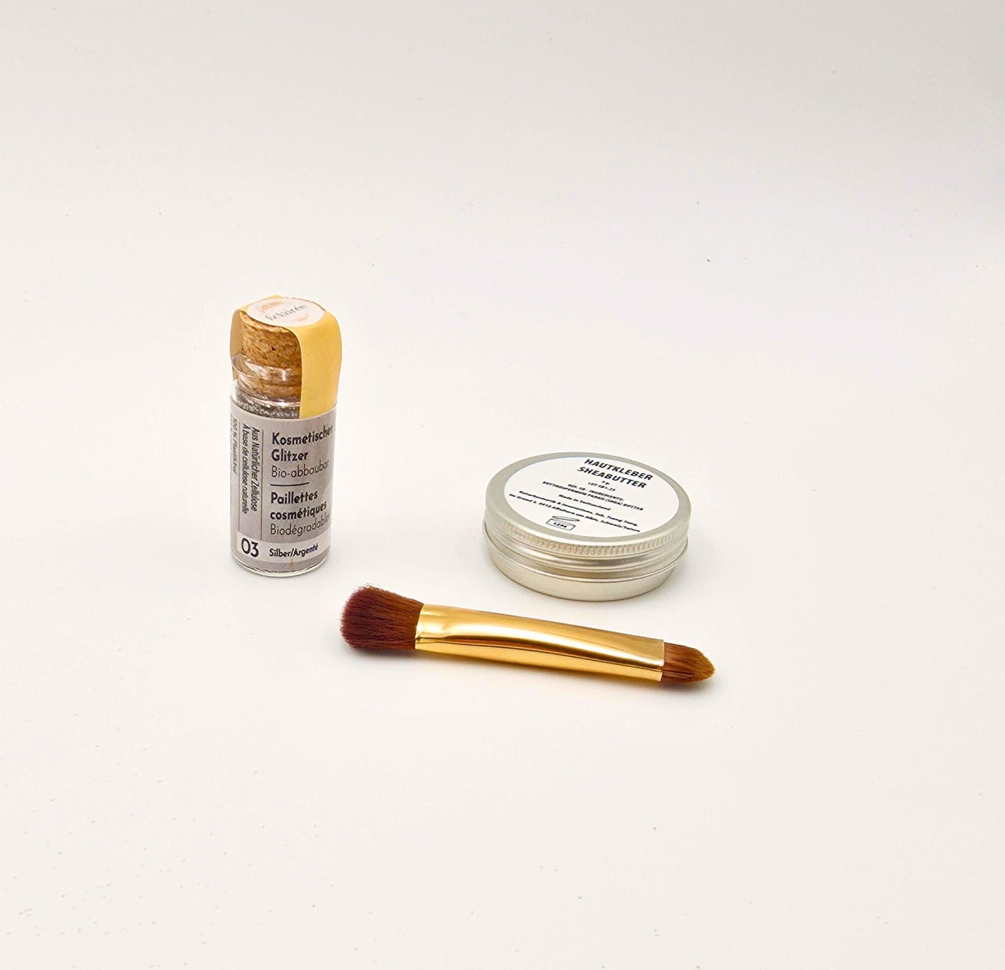 Set aus Shea-Butter, Pinsel und silbernem Bio-Glitzer in Glasflasche, sauber angeordnet für ein schimmerndes Beauty-Foto.