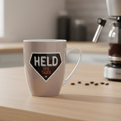 Grauer Porzellanbecher mit Aufdruck ''Held des Tages'' neben einer Kaffeemaschine – Geschenkidee für Kaffeeliebhaber