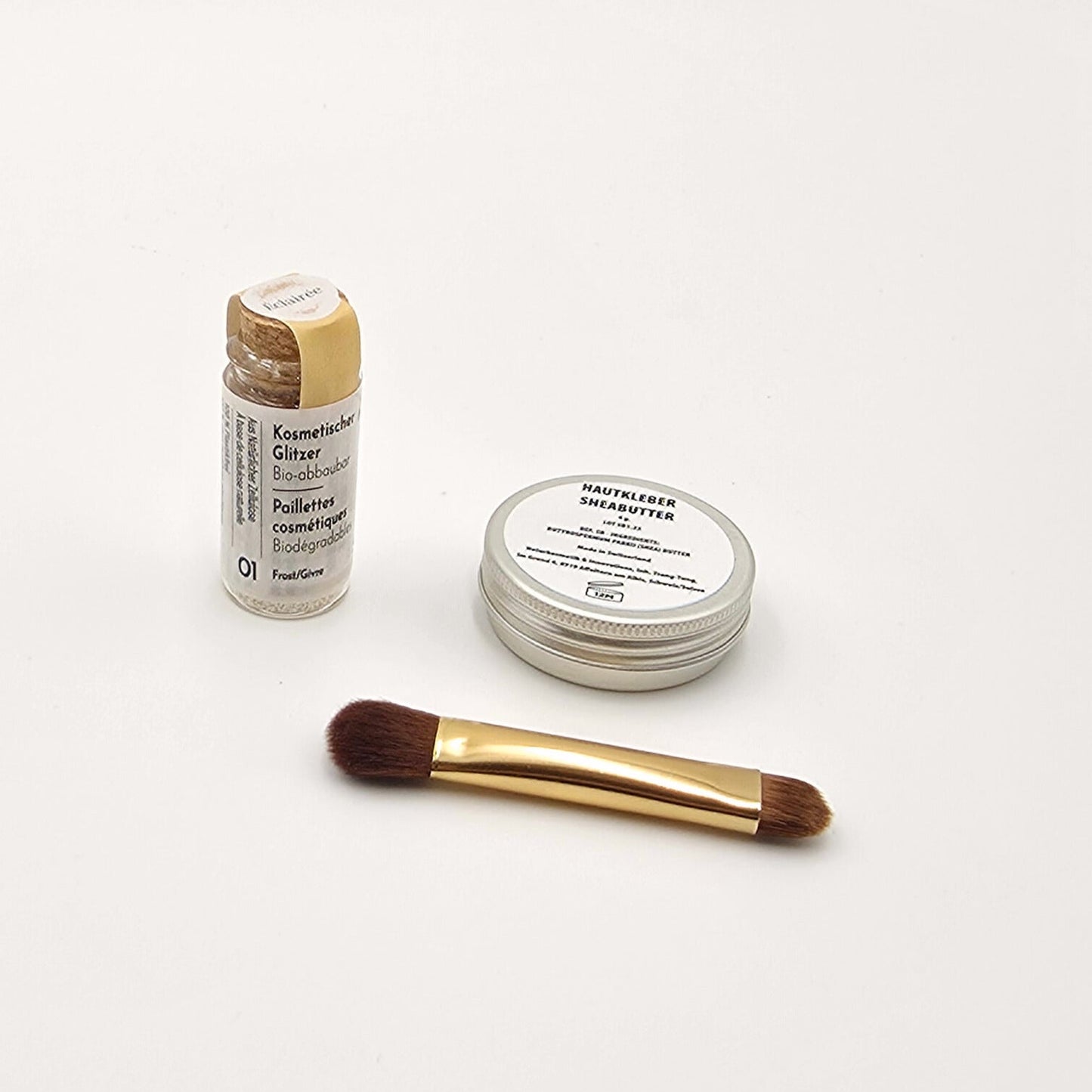 Shea-Butter-Dose, Pinsel und frostig-weiss schimmernder Bio-Glitzer in Glasflasche, minimalistisch präsentiert.