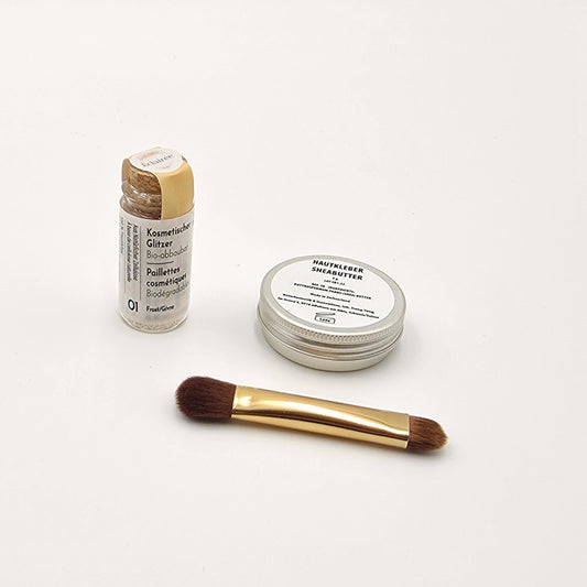 Shea-Butter-Dose, Pinsel und frostig-weiss schimmernder Bio-Glitzer in Glasflasche, minimalistisch präsentiert.