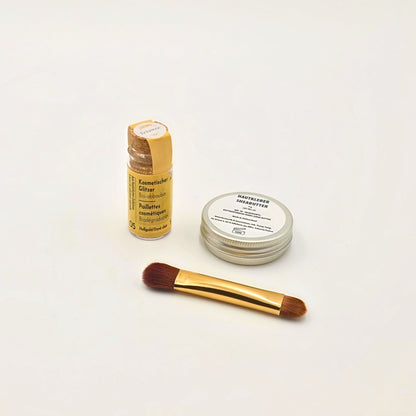 Shea-Butter-Dose, Auftragspinsel und Fläschchen mit hellgoldenem Bio-Glitzer, stilvoll arrangiert auf neutralem Hintergrund.