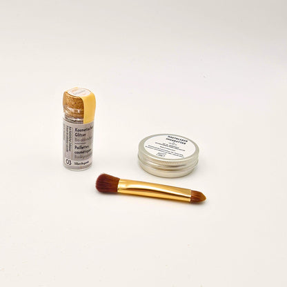 Set aus Shea-Butter, Pinsel und silbernem Bio-Glitzer in Glasflasche, sauber angeordnet für ein schimmerndes Beauty-Foto.