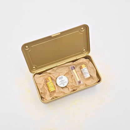 Offene goldene Geschenkbox, gefüllt mit Bio-Glitzer in Gold und Frost, Shea-Butter-Dose und feinem Pinsel, stilvoll arrangiert.