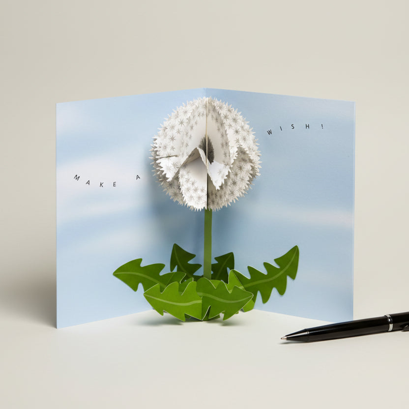 Geöffnete 3D Pop-up Grusskarte „Dandelion Wishes“ mit Pusteblumen-Motiv, blau und weiss mit Kugelschreiber als Grössenreferenz