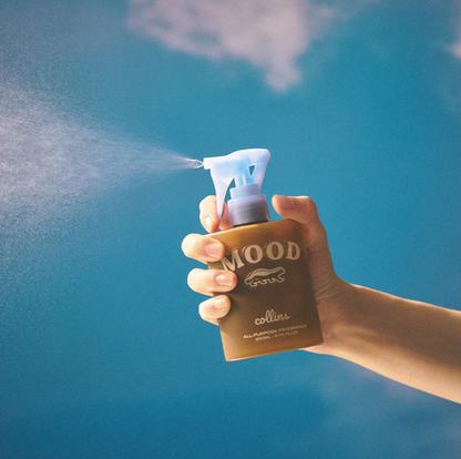 Eine Hand hält das braune Collins Mood Water Scottish Juniper Raumspray und versprüht feinen Duftnebel vor blauem Himmel – Erfrischendes Textilspray und Duftspray.