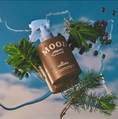 Braune Collins Mood Water Scottish Juniper Flasche liegend auf einer blauen, spiegelnden Fläche umgeben von grünen Nadelbaumzweigen – Holziger Raumduft für Zuhause.