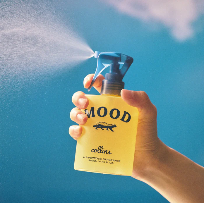 Eine Hand hält das gelbe Collins Mood Water Namhae Yuja Raumspray und versprüht feinen Duftnebel vor blauem Himmel – Erfrischendes Textilspray und Duftspray.