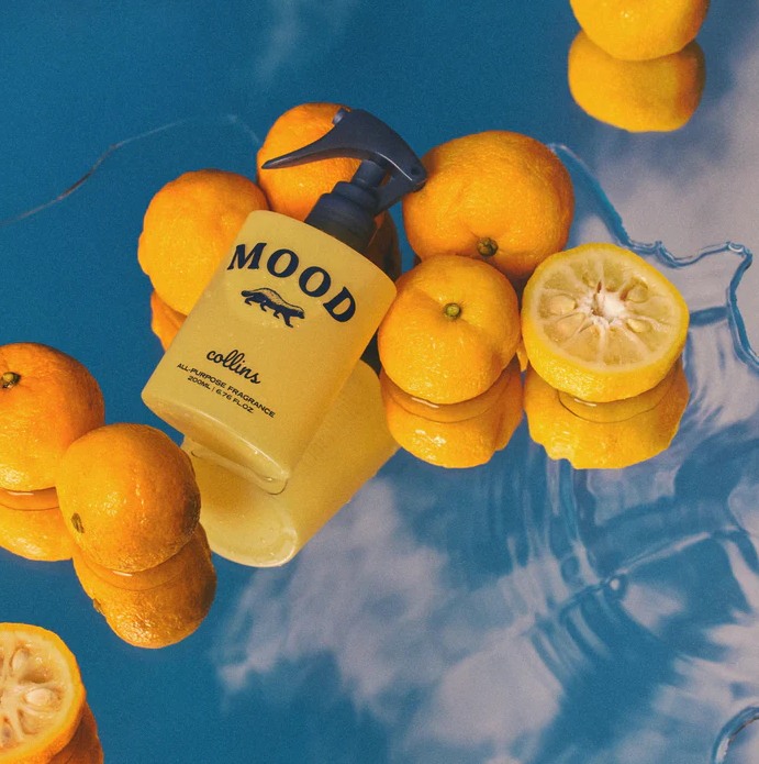 Gelbe Collins Mood Water Namhae Yuja Flasche liegend auf einer blauen, spiegelnden Fläche umgeben von halbierten und ganzen Yuzu-Früchten – Spritziger Raumduft.